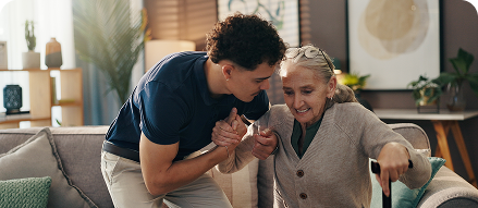 A young caregiver gently helping an elderly woman rise from a couch at home, both smiling during the interaction