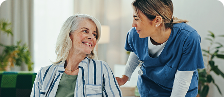 A smiling senior woman in a striped shirt sharing a warm moment with a caregiver in blue scrubs in a bright, plant-filled living room
