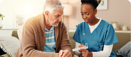 A home caregiver in blue scrubs reviewing medication or documents with an elderly man seated on a couch in a warmly lit living room