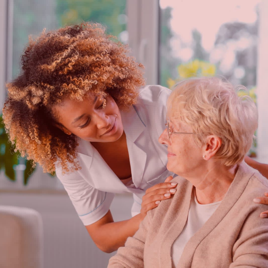 A smiling home caregiver in a white uniform gently placing her hand on an elderly woman's shoulder in a bright, plant-filled room